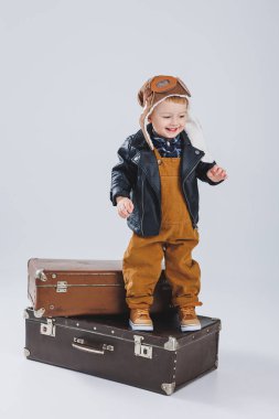 Deri ceketli ve kahverengi tulumlu küçük bir çocuk büyük bir bavulun üzerinde duruyor. Bavullu bir çocuk. Küçük gezgin..
