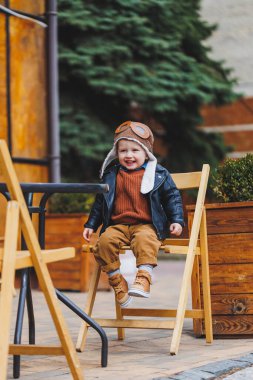 Üç yaşında, deri ceketli, kahverengi pantolonlu şık bir çocuk kafenin yanındaki terasta oturuyor. Modern çocuk. Çocuk modası. Mutlu çocuk