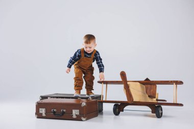 Kahverengi tulumlu neşeli bir çocuk bavulun üzerinde duruyor. Bavullu küçük bir gezgin. Tahta uçak, çocuk oyuncakları.