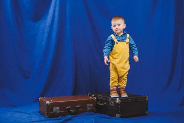 Ukraynalı 3 yaşındaki sarı tulumlu çocuk valizlerle oynuyor. Mavi arka planda modern bir çocuk. Mutlu çocuk.