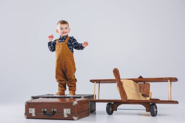 Kahverengi tulumlu neşeli bir çocuk bavulun üzerinde duruyor. Bavullu küçük bir gezgin. Tahta uçak, çocuk oyuncakları.