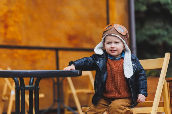 Üç yaşında, deri ceketli, kahverengi pantolonlu şık bir çocuk kafenin yanındaki terasta oturuyor. Modern çocuk. Çocuk modası. Mutlu çocuk
