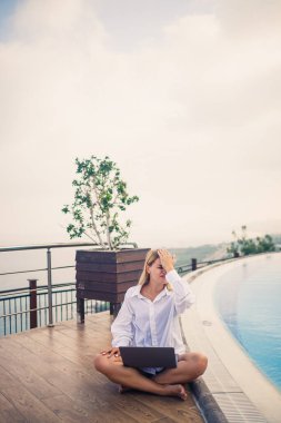 Güzel ve çekici genç bir kadın büyük bir havuzun yanında oturuyor ve bir dizüstü bilgisayarda çalışıyor. Tatildeyken uzak çalışma.