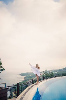 Siyah mayolu güzel bir kadın açık havuz kenarında deniz manzaralı poz veriyor.