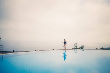 Siyah mayolu güzel bir kadın açık havuz kenarında deniz manzaralı poz veriyor.