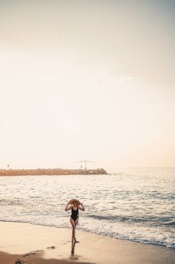 Siyah mayo ve gözlüklü şapkalı güzel bir kadın gün batımında Türkiye sahilinde yürüyor. Deniz eğlencesi konsepti. Seçici odak