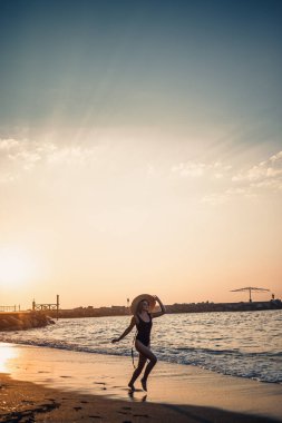 Siyah mayo ve gözlüklü şapkalı güzel bir kadın gün batımında kumsalda yürüyor. Deniz eğlencesi konsepti. Seçici odak
