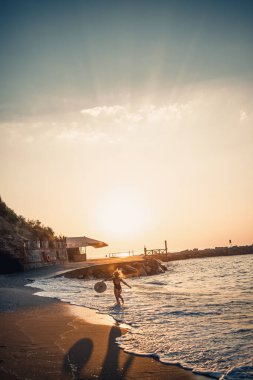 Siyah mayo ve gözlüklü şapkalı güzel bir kadın gün batımında kumsalda yürüyor. Deniz eğlencesi konsepti. Seçici odak