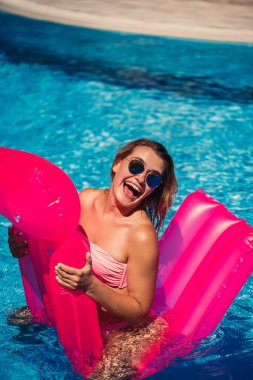 Yüzünde mayo olan güneş gözlüklü seksi kadın havuzda pembe şişme bir şiltenin üzerinde yatıyor. Sıcak ve güneşli bir yaz gününde havuz kenarında rahatla. Tatil konsepti. Seçici odak