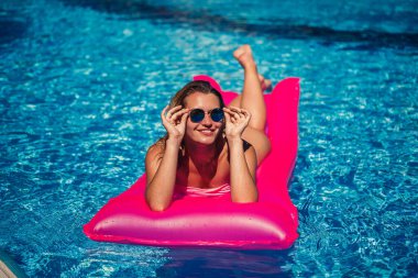 Havuzdaki pembe şişme yatakta mayo giymiş seksi bir kadın yatıyor. Sıcak ve güneşli bir yaz gününde havuz kenarında rahatla. Tatil kavramı