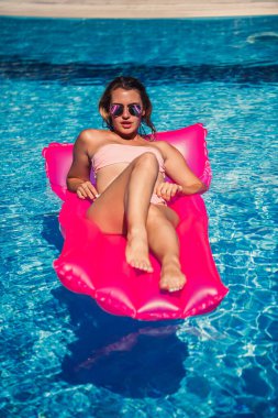 Seksi kadın model dinleniyor ve havuzda şiltede güneşleniyor. Pembe bikinili bir kadın şişme pembe bir yatakta yüzüyor. spf ve güneş kremi