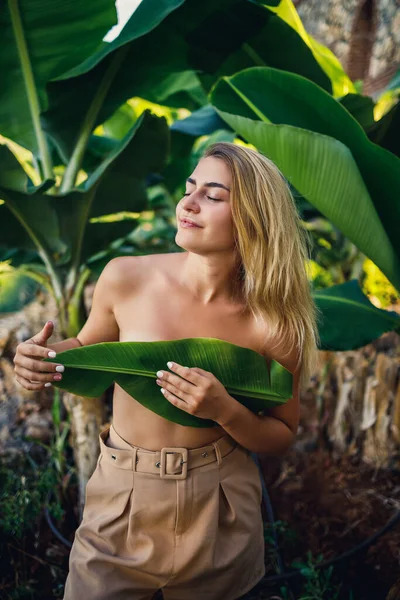 美しい若い女性がバナナの木の近くの美しいポーズに立って 女の子はバナナのプランテーションで彼女の胸を覆います — ストック写真