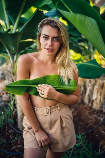美しい若い女性はバナナの木の近くの美しいポーズに立っています 彼女の女の子はバナナのプランテーションに葉で彼女の胸をカバーします — ストック写真