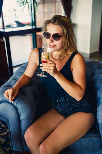 美しい若い女性が休暇中にシャンパンのガラスとホテルのロビーで柔らかいアームチェアに座っています 顔にはサングラスと笑顔をかけている 休暇の概念 — ストック写真
