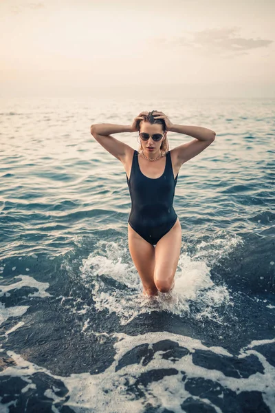 Güneş gözlüklü çekici genç bir kadın gün batımında siyah bir mayoyla denizden çıkıyor. Seçici odak