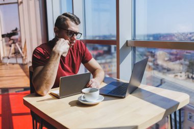 Modern bir dizüstü bilgisayarla uzaktan çalışan, mola sırasında masaya oturup kameraya gülümseyen, vizyon düzeltme gözlüklü mutlu bir insan programcı portresi.