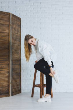 Siyah pantolonlu genç bir kadın ve beyaz gömlekli beyaz tuğlalı bir duvara karşı şık, modern bir kız, beyaz deri ayakkabılı.