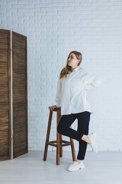 Siyah pantolonlu genç bir kadın ve beyaz gömlekli beyaz tuğlalı bir duvara karşı şık, modern bir kız, beyaz deri ayakkabılı.