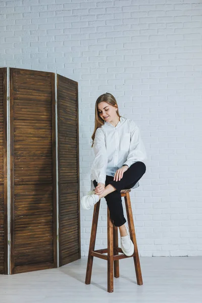 Güzel bir kadın beyaz pamuklu gömlek ve siyah pantolon giyiyor. Neşeli duygusal kız beyaz bir duvara karşı tahta bir sandalyede oturuyor..