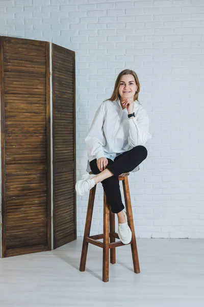 Güzel bir kadın beyaz pamuklu gömlek ve siyah pantolon giyiyor. Neşeli duygusal kız beyaz bir duvara karşı tahta bir sandalyede oturuyor..