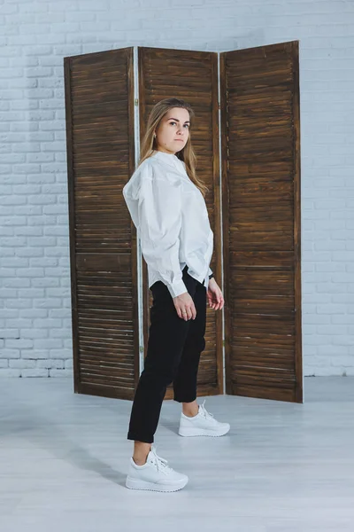 Güzel, şık bir kadın beyaz pamuklu gömlek ve siyah pantolon giyiyor. Beyaz spor ayakkabılı neşeli duygusal kız.