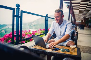 Başarılı bir erkek işadamı dizüstü bilgisayarla masada oturuyor ve tatilde. Tatil ve uzaktan kumanda. Adam terasta panoramik manzaralı bir restoranda oturuyor.