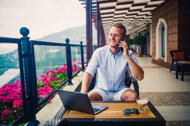 Beyaz gömlekli ve şortlu genç yakışıklı bir işadamı masada dizüstü bilgisayarla oturuyor ve telefonla konuşuyor. Dinlenirken çalış