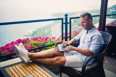 Beyaz gömlekli ve şortlu genç yakışıklı iş adamı masadaki bir kafede dizüstü bilgisayarla oturuyor. Dinlenirken çalış. Seçici odak