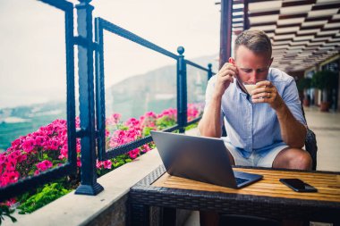 Serbest yazar konsepti. Başarılı genç adam, iş adamı dizüstü bilgisayarda çalışıyor, terasta oturuyor kahve içiyor ve telefonda konuşuyor..