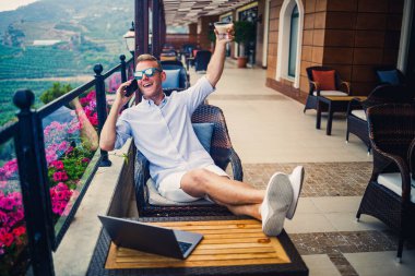 Serbest yazar konsepti. Başarılı genç adam, iş adamı dizüstü bilgisayarda çalışıyor, terasta oturuyor kahve içiyor ve telefonda konuşuyor..