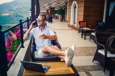 Serbest yazar konsepti. Başarılı genç adam, iş adamı dizüstü bilgisayarda çalışıyor, terasta oturuyor kahve içiyor ve telefonda konuşuyor..