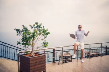 Dizüstü bilgisayarla çalışan yakışıklı ve başarılı genç iş adamı Akdeniz 'e bakıyor. O bir gömlek ve beyaz şort giyiyor. Tatilde uzaktan çalışma. Tatil kavramı