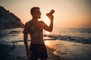 Genç erkek fotoğrafçı gün batımında kıyıda dururken denizin fotoğraflarını çeker. Deniz batımı. Seçici odaklanma. Turist fotoğrafları deniz manzarası