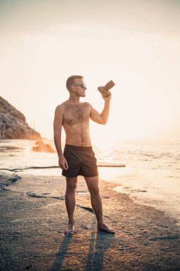Genç erkek fotoğrafçı gün batımında kıyıda dururken denizin fotoğraflarını çeker. Deniz batımı. Seçici odaklanma. Turist fotoğrafları deniz manzarası