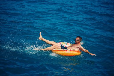 Genç bir adam mavi suyla denizde şişirilebilir bir halka üzerinde yüzüyor. Güneşli bir günde güzel bir tatil..