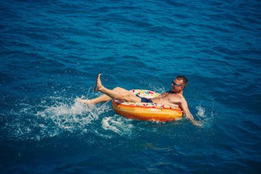 Genç bir adam güneşli bir günde şişme bir yüzükle açık denizde yüzer. Yaz tatili, tatildeki turist.