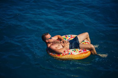 Genç bir adam güneşli bir günde şişme bir yüzükle açık denizde yüzer. Yaz tatili, tatildeki turist.