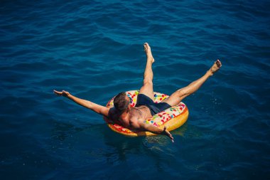 Genç bir adam mavi suyla denizde şişirilebilir bir halka üzerinde yüzüyor. Güneşli bir günde güzel bir tatil..
