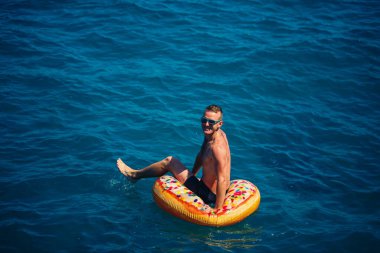 Genç bir adam mavi suyla denizde şişirilebilir bir halka üzerinde yüzüyor. Güneşli bir günde güzel bir tatil. Tatil konsepti, üst görünüm.