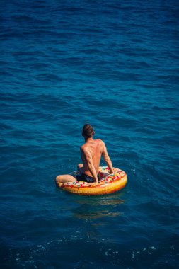 Genç bir adam mavi suyla denizde şişirilebilir bir halka üzerinde yüzüyor. Güneşli bir günde güzel bir tatil. Tatil konsepti, üst görünüm.