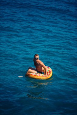 Genç bir adam mavi suyla denizde şişirilebilir bir halka üzerinde yüzüyor. Güneşli bir günde güzel bir tatil. Tatil konsepti, üst görünüm.
