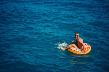 Genç bir adam mavi suyla denizde şişirilebilir bir halka üzerinde yüzüyor. Güneşli bir günde güzel bir tatil. Tatil konsepti, üst görünüm.