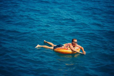Genç bir adam mavi suyla denizde şişirilebilir bir halka üzerinde yüzüyor. Güneşli bir günde güzel bir tatil..