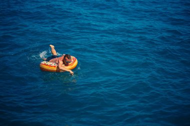 Genç bir adam mavi suyla denizde şişirilebilir bir halka üzerinde yüzüyor. Güneşli bir günde güzel bir tatil..