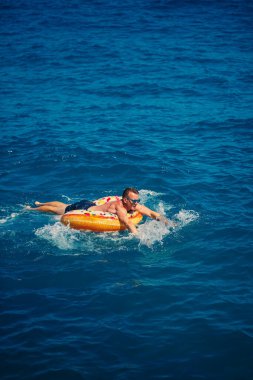 Bir adam şişme kauçuk bir çemberin üzerinde uzanır ve açık güneşli bir yaz gününde mavi denizde yüzer.