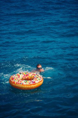 Genç bir adam mavi suyla denizde şişirilebilir bir halka üzerinde yüzüyor. Güneşli bir günde güzel bir tatil..