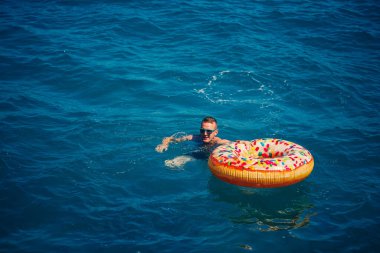 Genç adam güneşli bir günde denizde şişme bir yüzükte dinleniyor ve yüzüyor.