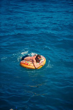 Genç adam güneşli bir günde denizde şişme bir yüzükte dinleniyor ve yüzüyor.