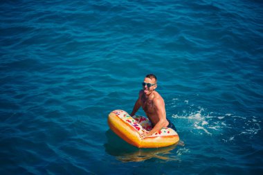 Genç adam güneşli bir günde denizde şişme bir yüzükte dinleniyor ve yüzüyor.