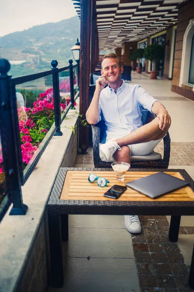 Beyaz gömlekli ve şortlu genç yakışıklı iş adamı masadaki bir kafede dizüstü bilgisayarla oturuyor. Dinlenirken çalış